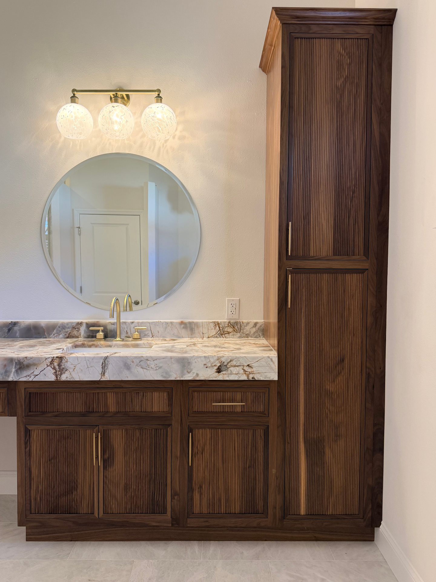 Walnut linen tower with fluted door fronts and brushed brass pulls beside a round mirror and globe sconces